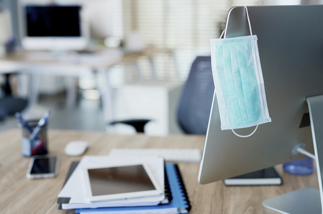 Disposable face mask hangs on the side of a computer screen on an employee's desk