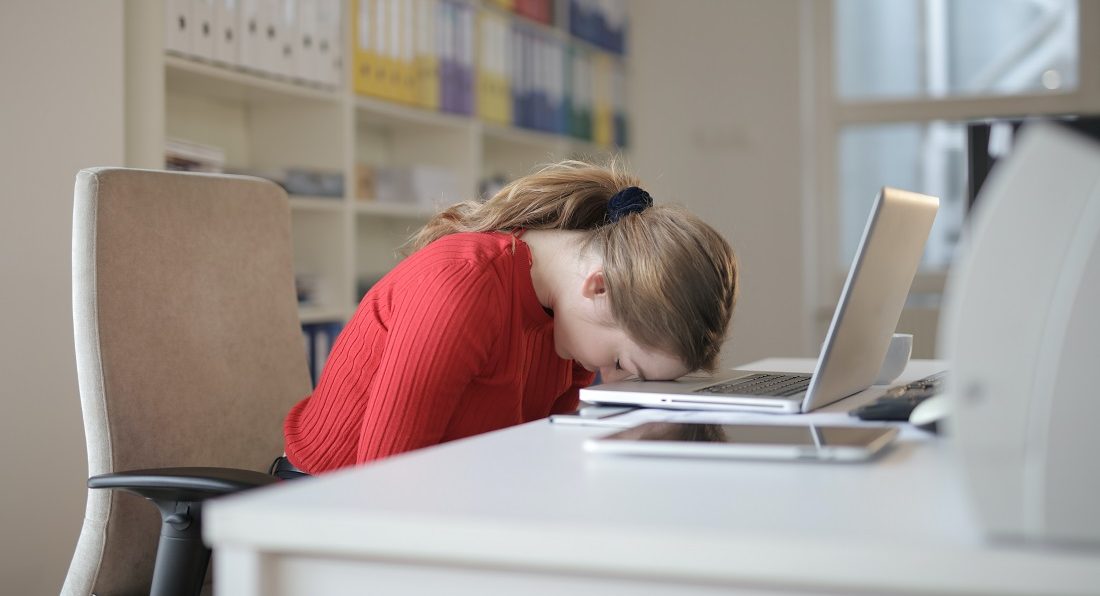 woman in a red shirt puts her head on her office desk and looks frustrated