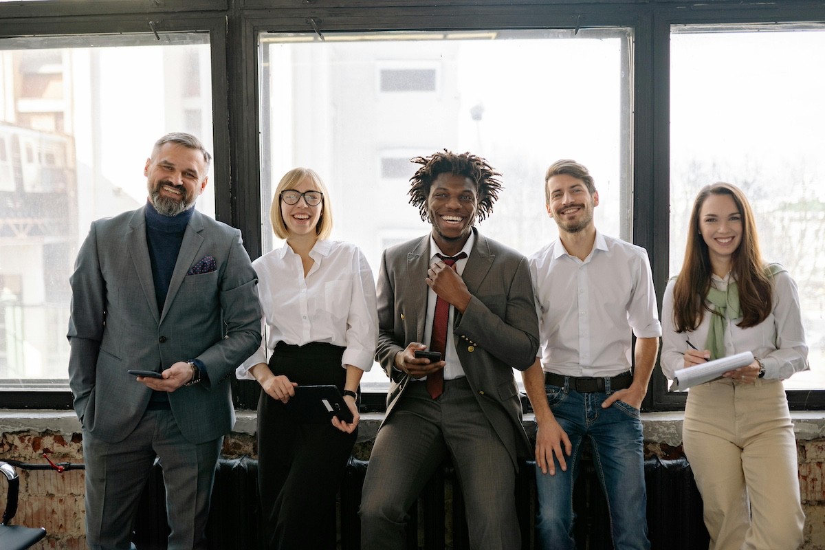 five employees dressed professionally and smiling sit next to the window in their office