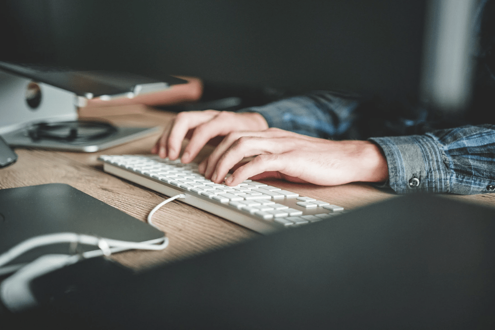 Person typing on their keyboard at their desk wearing a long-sleeved plaid shirt