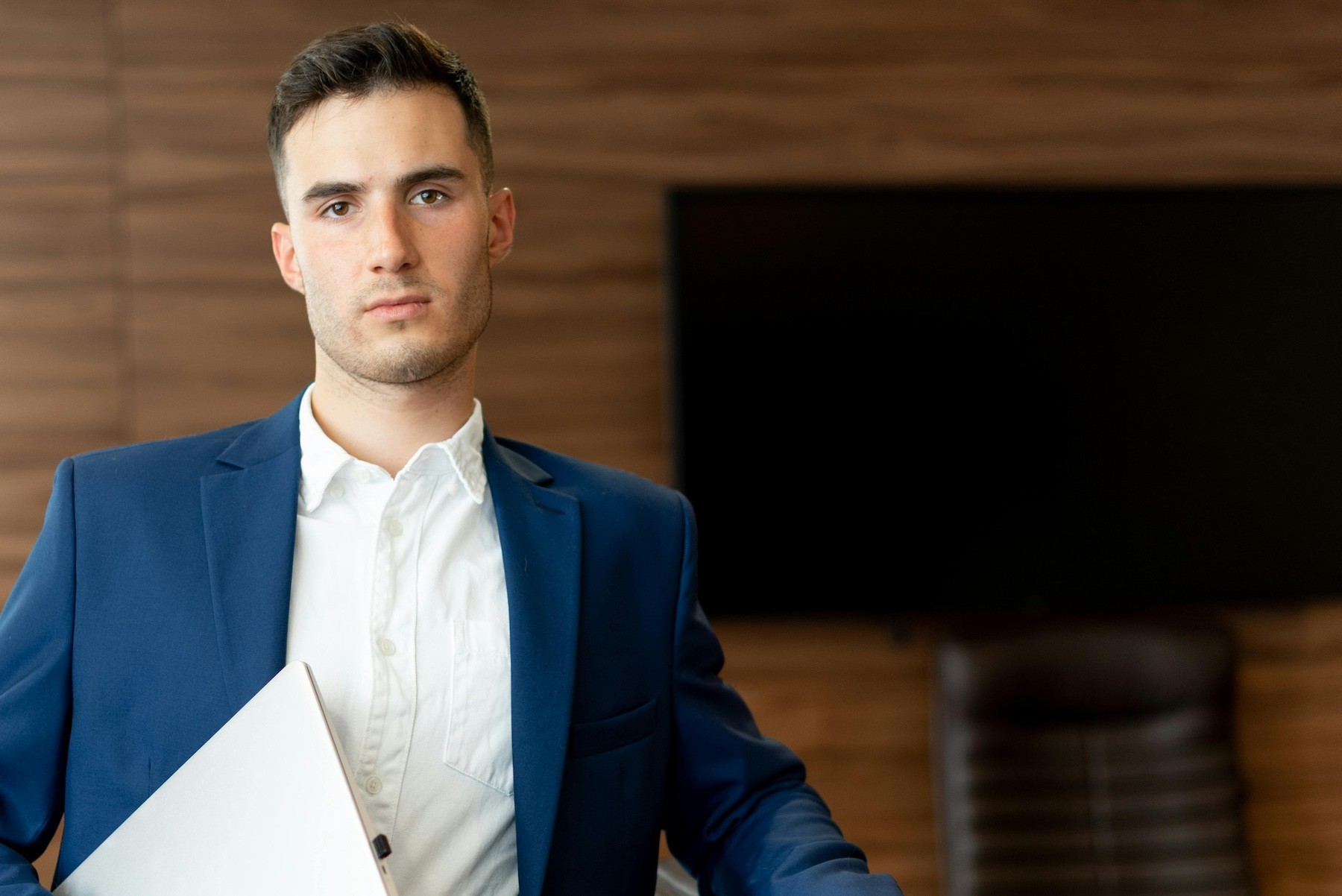 young working professional man in a blue suit and white collard shirt sits holding his laptop looking complacent