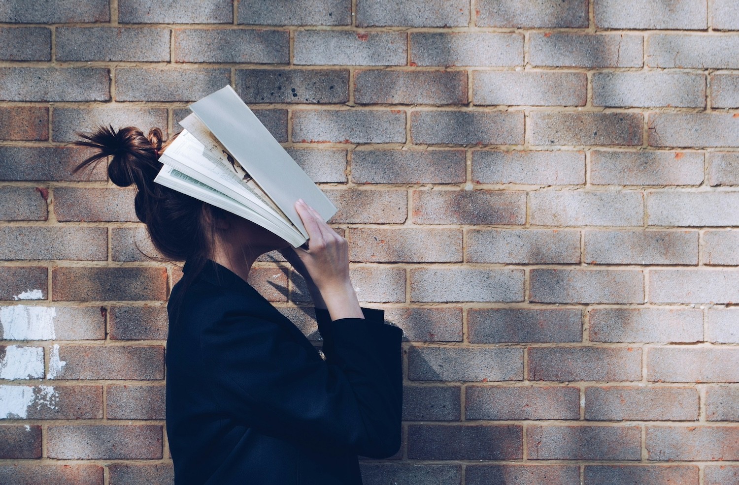 woman covering her face with white book standing in front of a brick wall