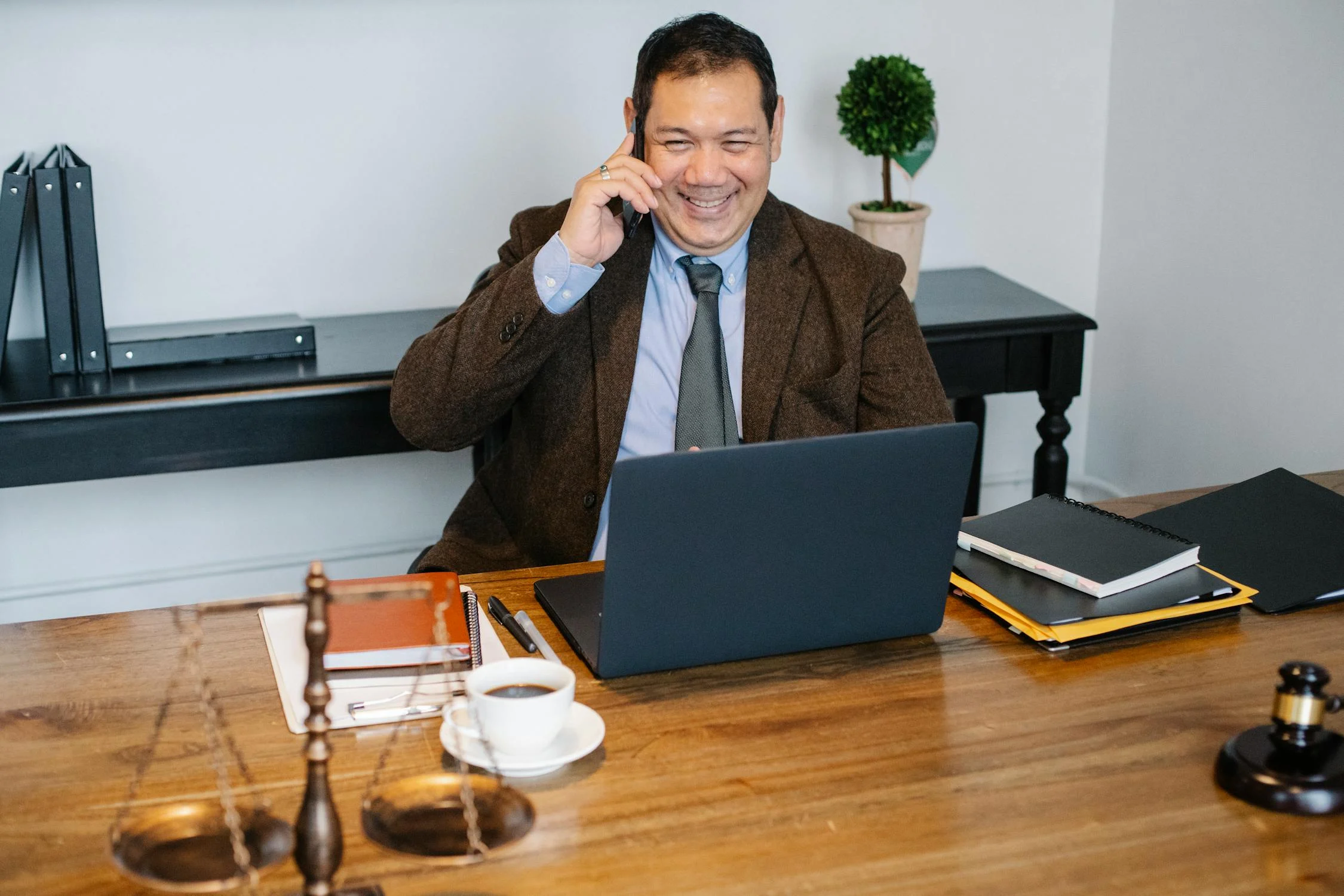 Professional man makes a phone call as he sits at his desk containing his office supplies and a lady justice statue.